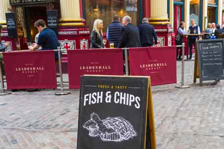 Leadenhall Market in London, fish and chips sign from the market in the front and people are eating and drinking in Leadenhall at the back